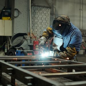 Young male welder in protective mask and gloves welding metallic structure or huge part of industrial machine in workshop