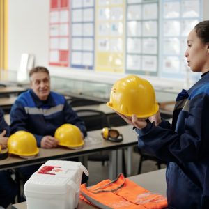 young-pretty-female-worker-uniform-standing-front-her-male-colleagues-while-describing-purpose-usage-protective-helmet